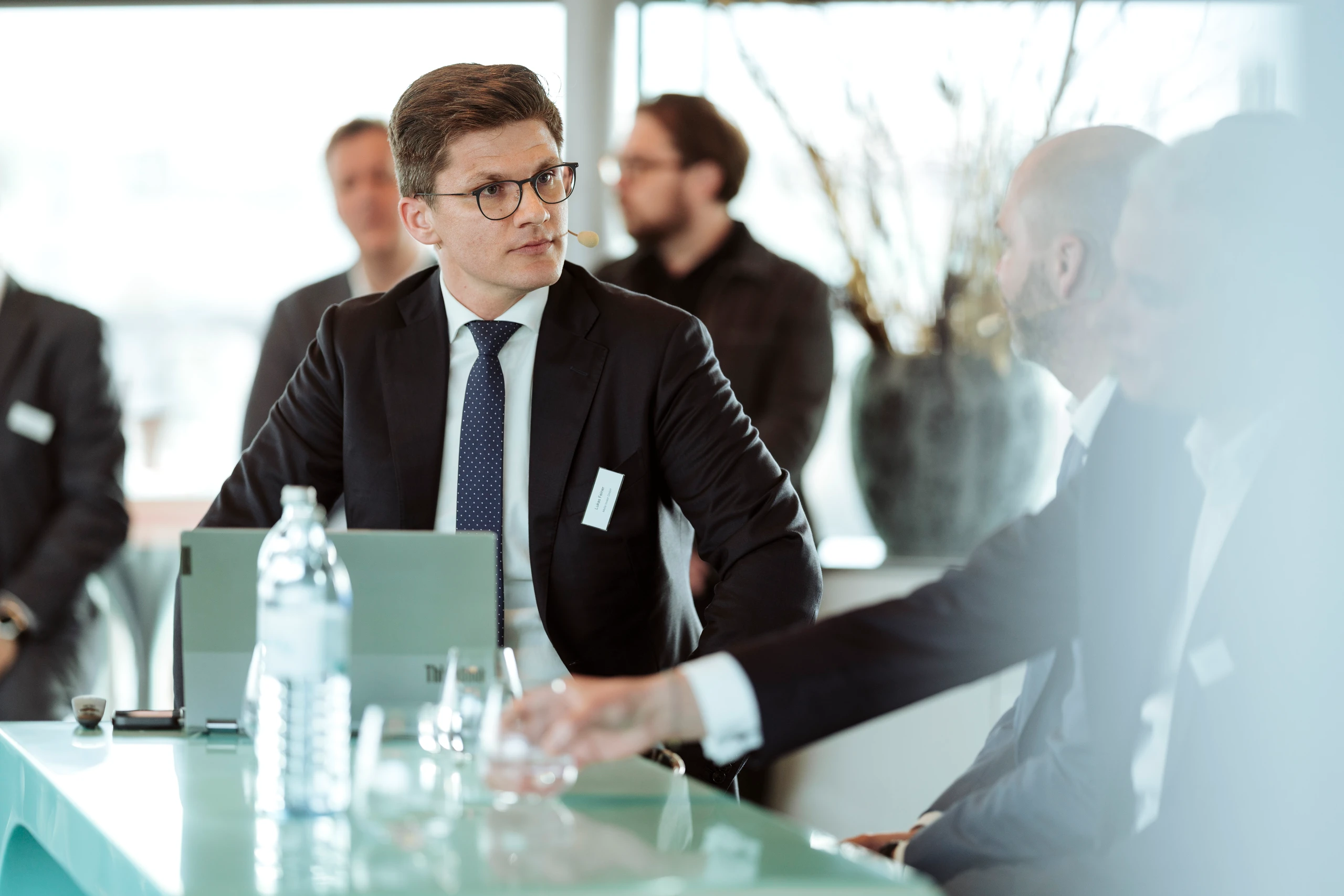 Mann im Anzug bei einem Business-Meeting.  Konzentrierter Blick, Laptop und Wasserflasche auf dem Tisch.
