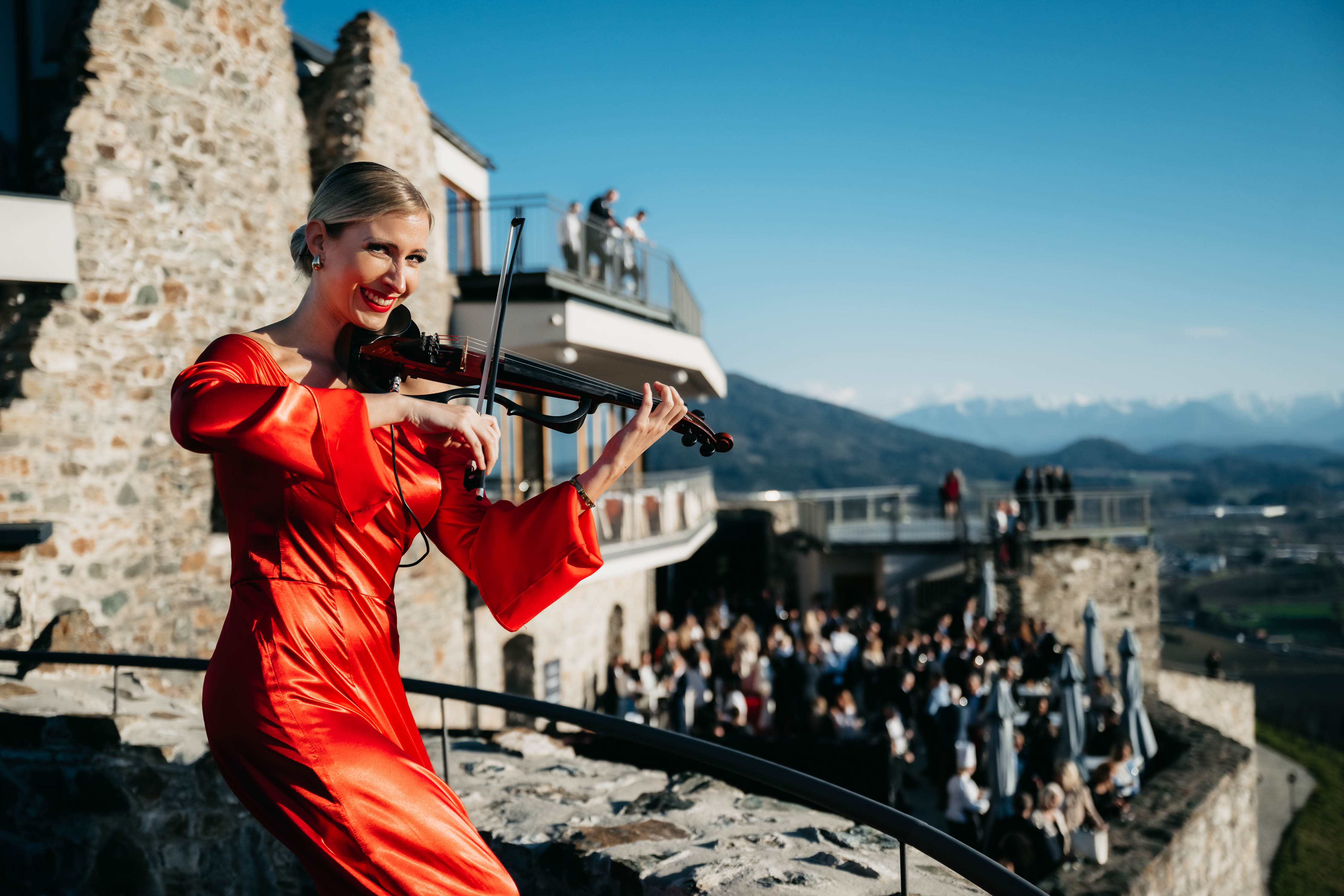 Geigenspielerin in rotem Kleid spielt auf einer Burg vor vielen Zuschauern und Bergen. Foto von MATIC KREMZAR.