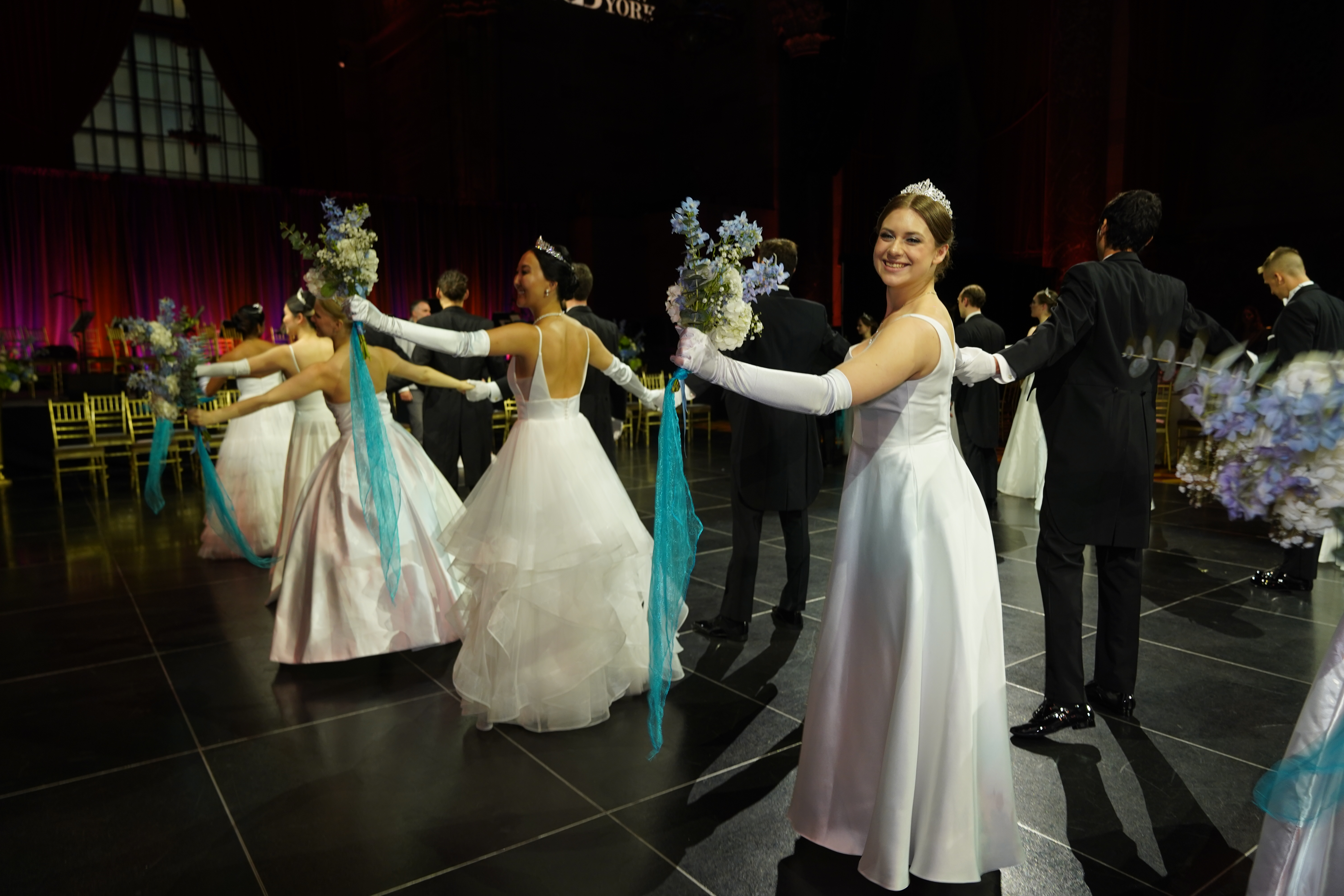 Junge Frauen in eleganten weißen Ballkleidern bei einem formellen Ball. Sie halten Blumensträuße und tragen weiße Handschuhe.