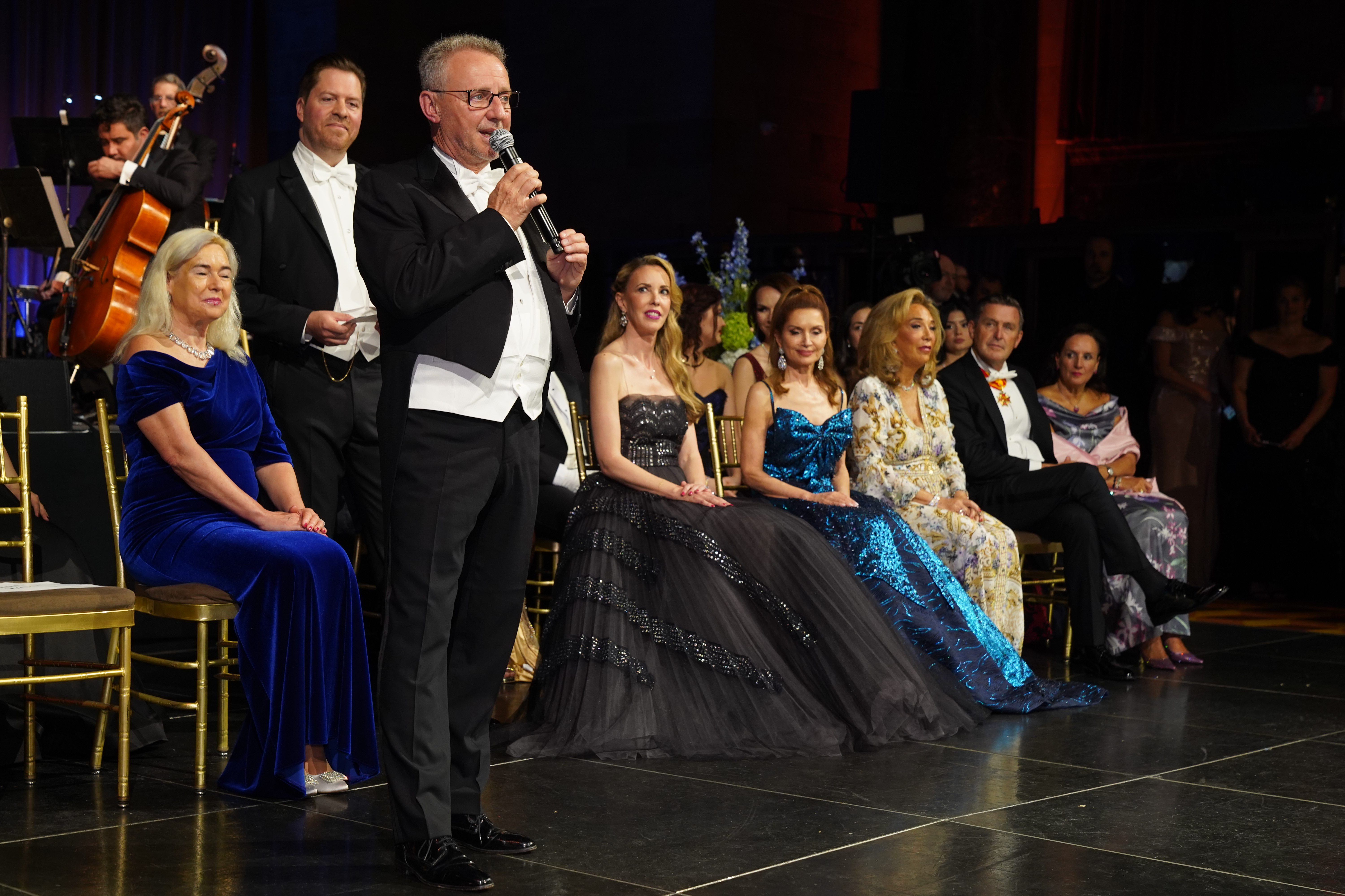 Mann in Smoking hält Rede auf Gala, Publikum in Abendkleidung, Orchester im Hintergrund.