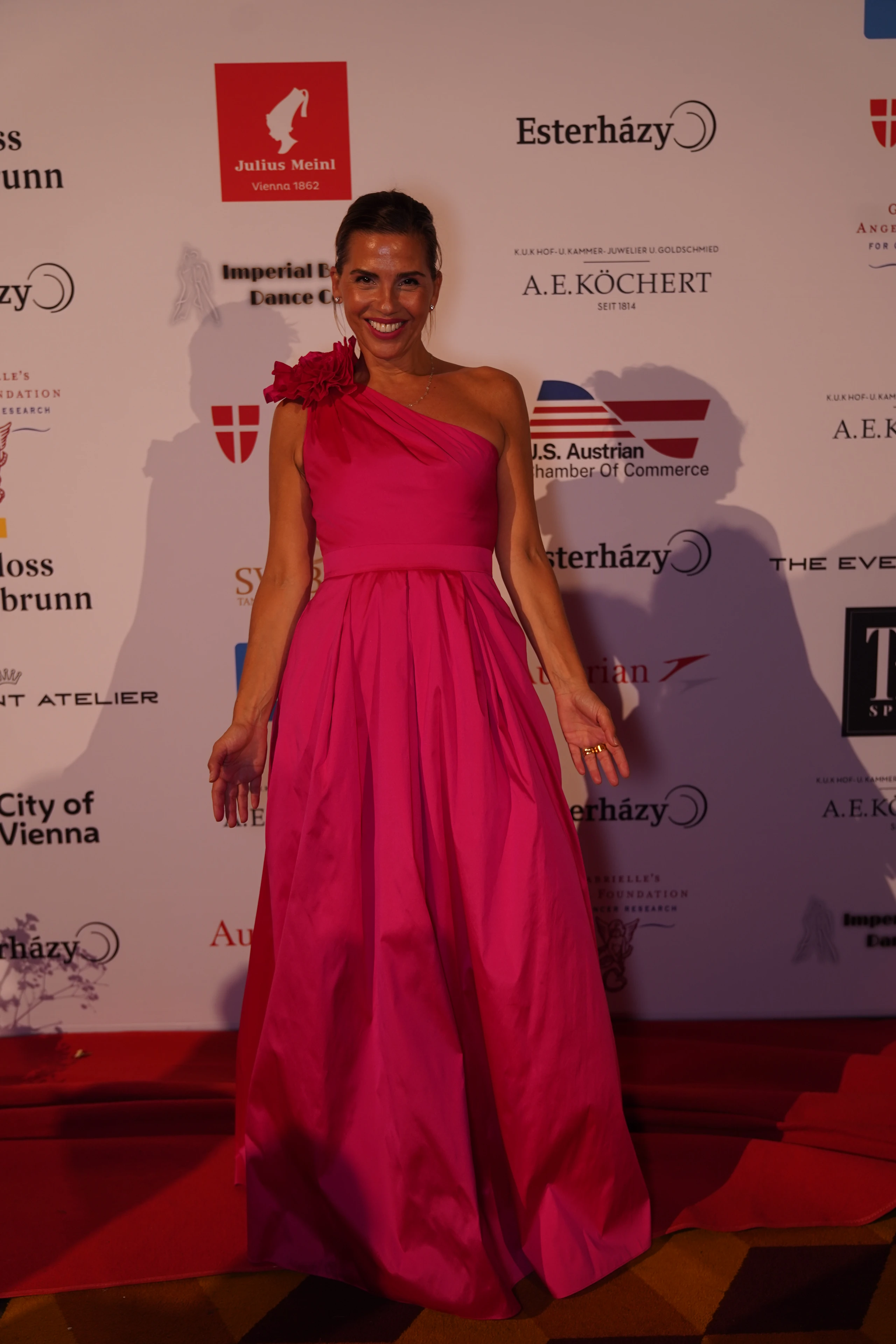 Frau in rosafarbenem Abendkleid auf Gala-Event. Bodenlanges Kleid mit Blütenapplikation.  Hintergrund mit Sponsorenlogos.