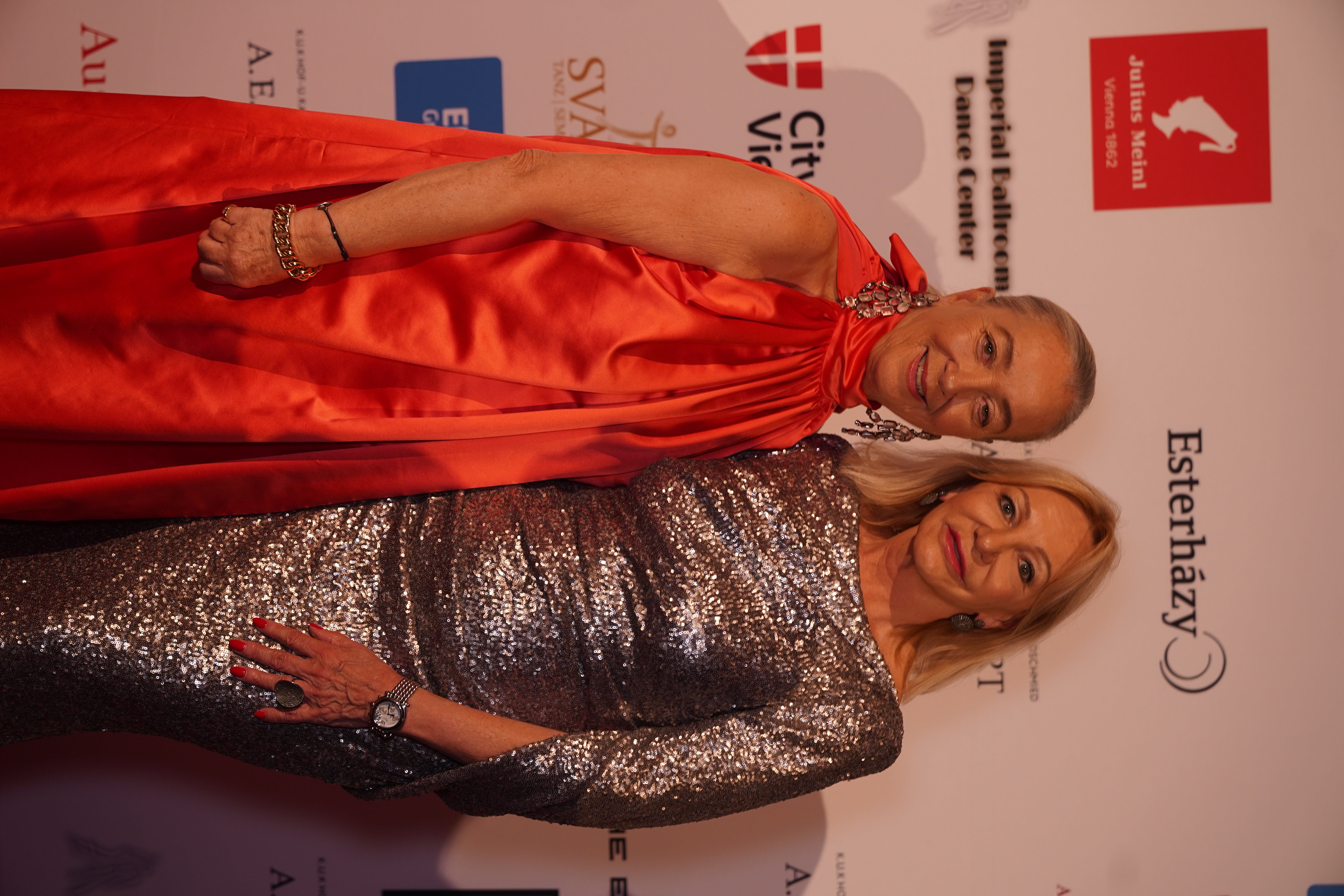 Zwei elegant gekleidete Frauen bei einer Gala in roten und silbernen Kleidern.