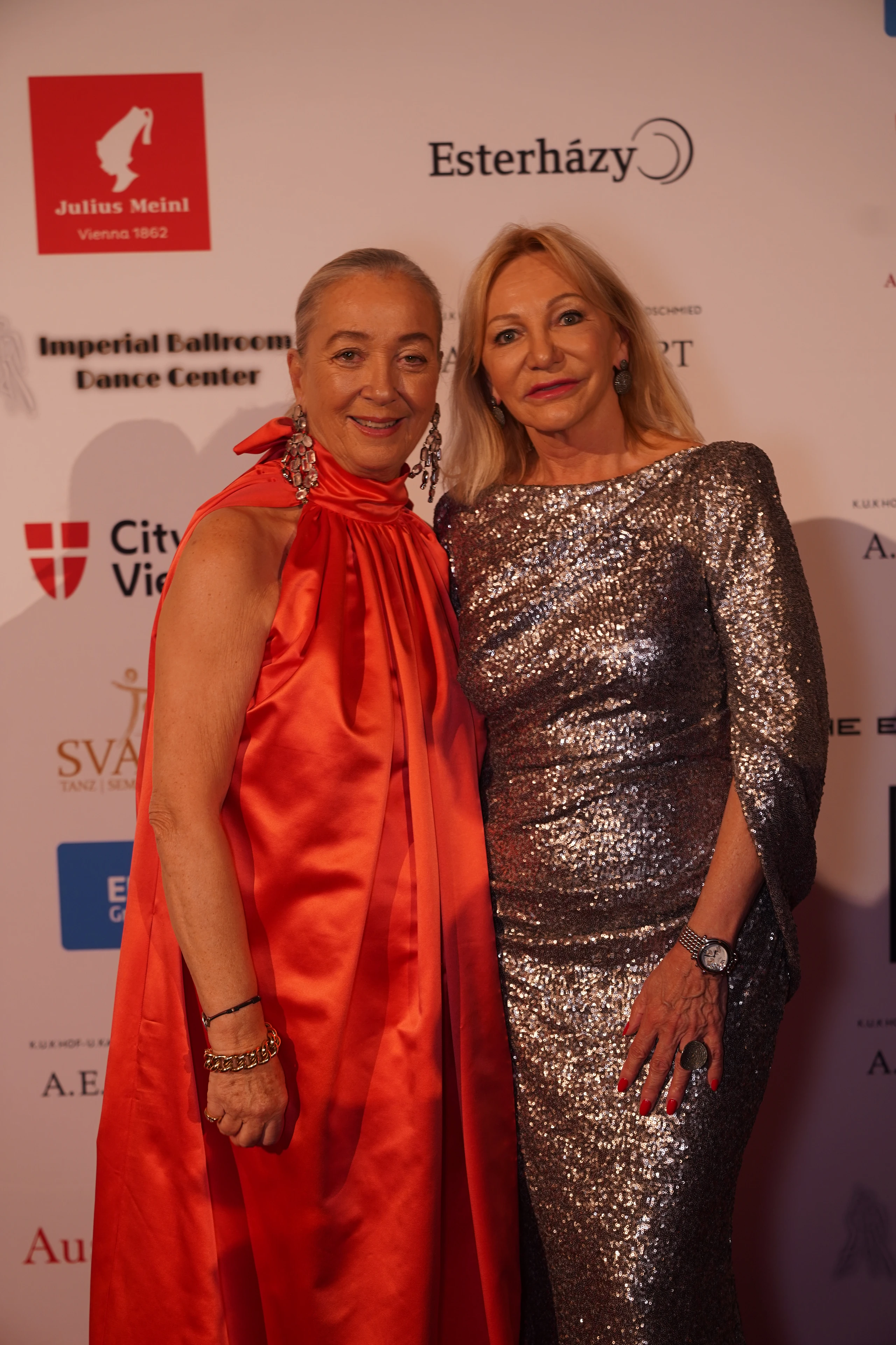 Zwei elegant gekleidete Frauen bei einer Gala in roten und silbernen Kleidern.