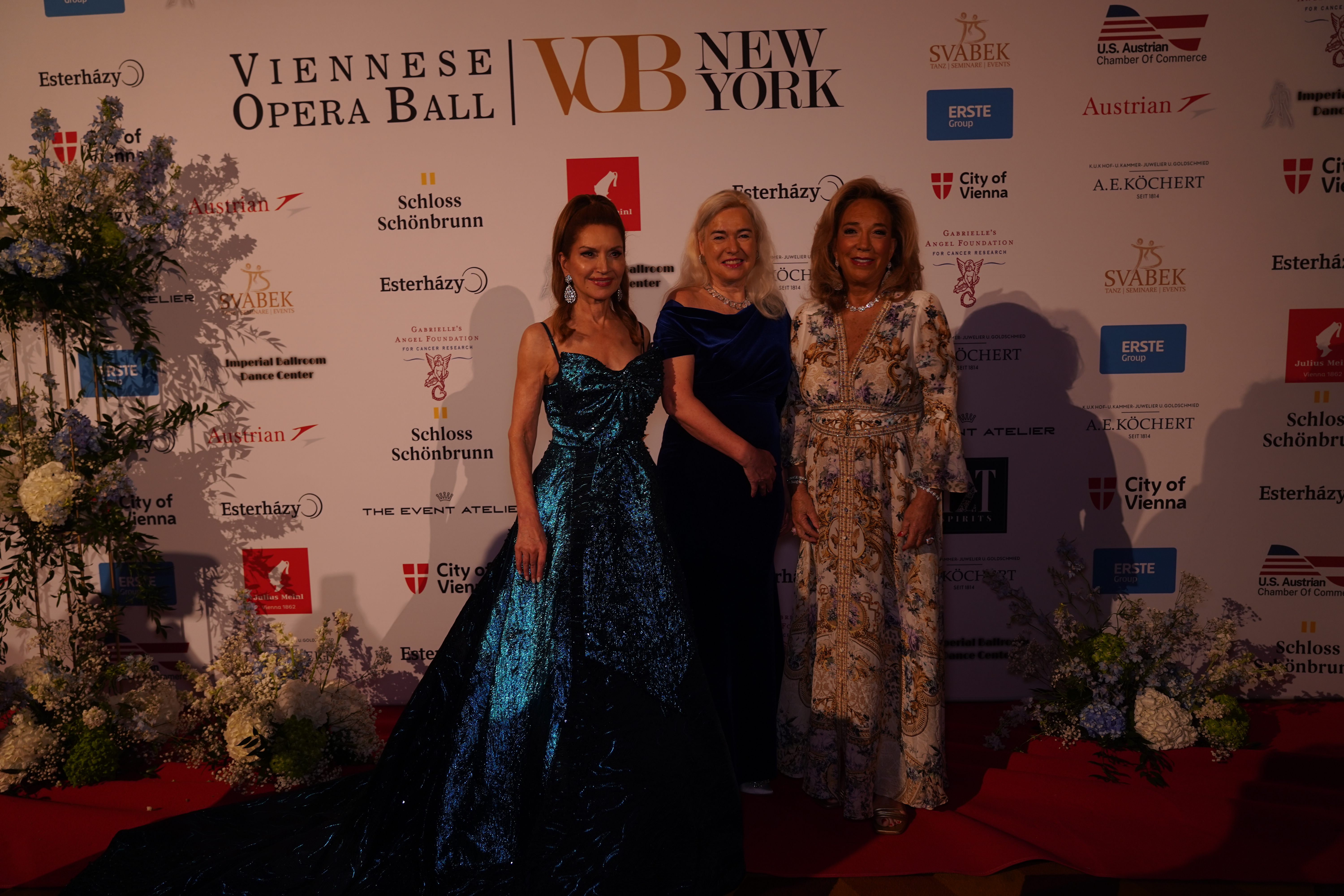 Drei Damen in Abendkleidern beim Wiener Opernball in New York.  Sponsorenlogos im Hintergrund.