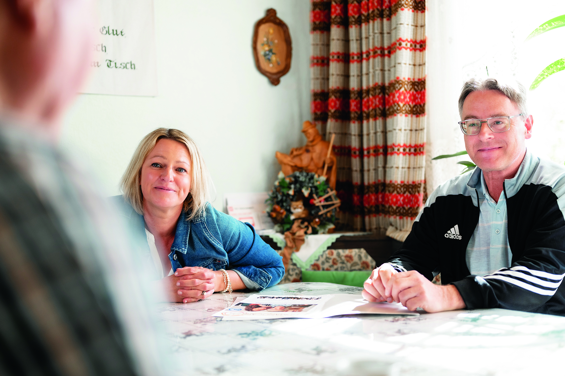 Entspanntes Paar unterhält sich an einem Küchentisch. Frau in Jeansjacke, Mann in Adidasjacke.  Erstellt mit Color Factory 8.1
