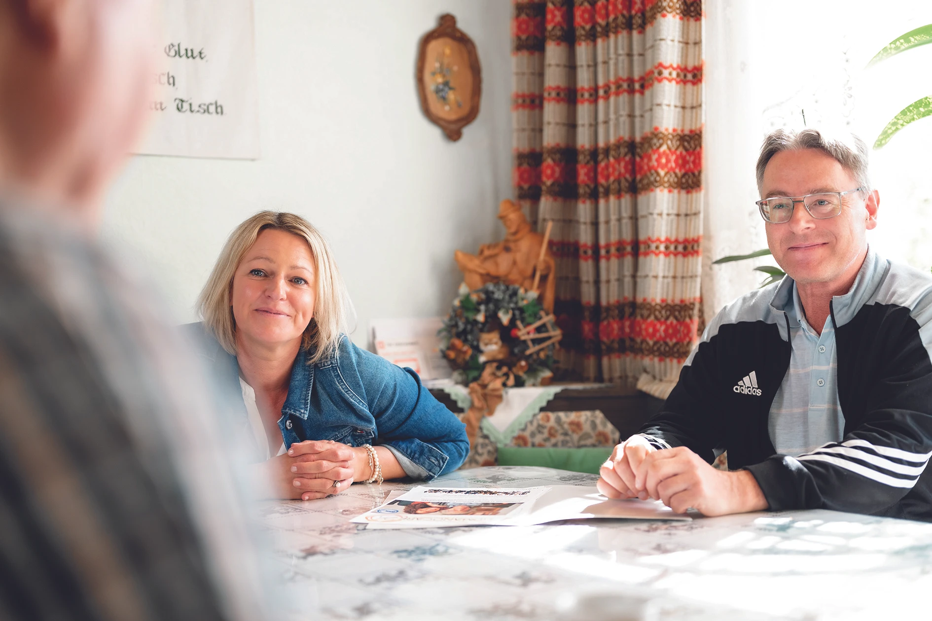 © Rene Knabl Entspanntes Paar unterhält sich an einem Küchentisch. Frau in Jeansjacke, Mann in Adidasjacke.  Erstellt mit Color Factory 8.1